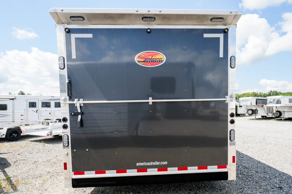 Back of a dark trailer with a Sundowner logo and a metal support bar across the door, parked in a lot with other trailers visible in the background.