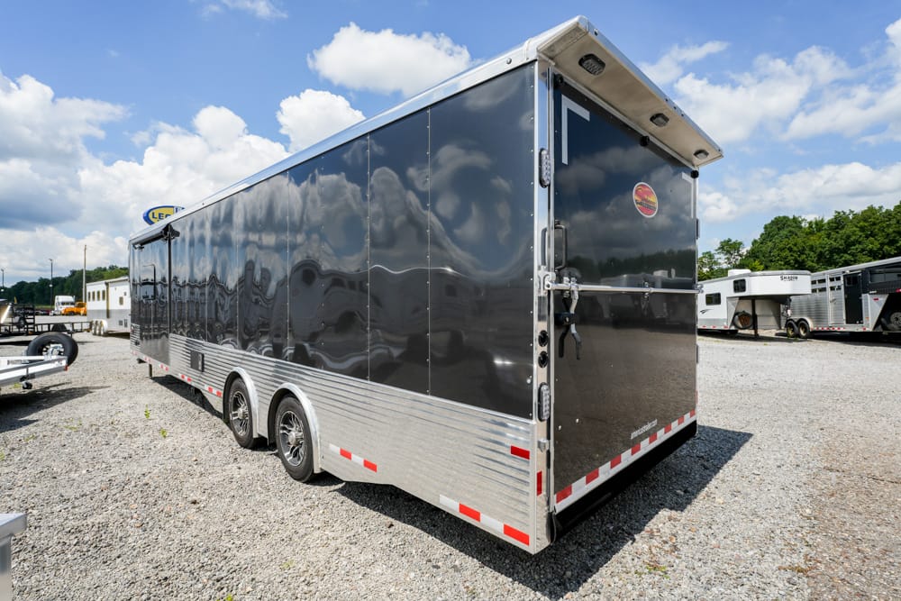 Black enclosed cargo trailer with two axles and aluminum trim parked on a gravel lot under a blue sky.