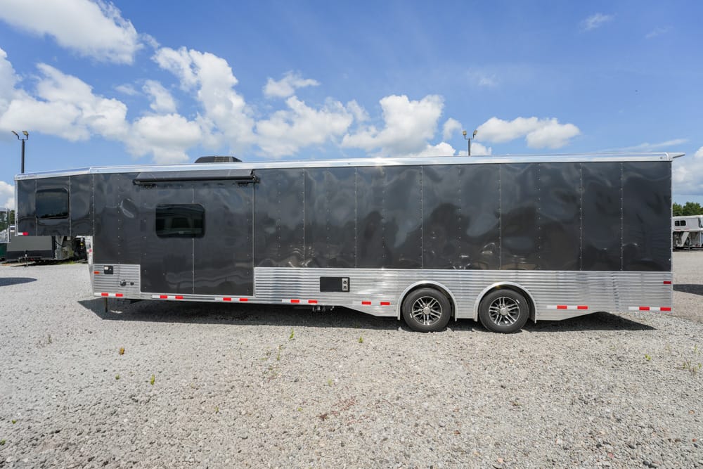 Large dark gray enclosed trailer with silver lower siding and two axles parked on a gravel lot under a partly cloudy blue sky.