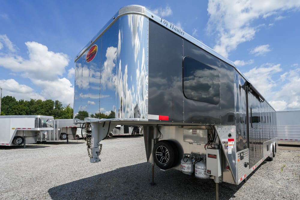Shiny dark gray toy hauler trailer with a metal front hitch, spare tire, and propane tanks on a gravel lot under a blue sky with clouds.