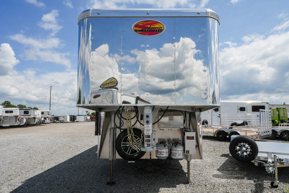 Polished aluminum gooseneck trailer front with Sundowner Trailers logo, parked in a gravel lot among other trailers; propane tanks and hookup hardware visible underneath, clear blue sky with clouds.
