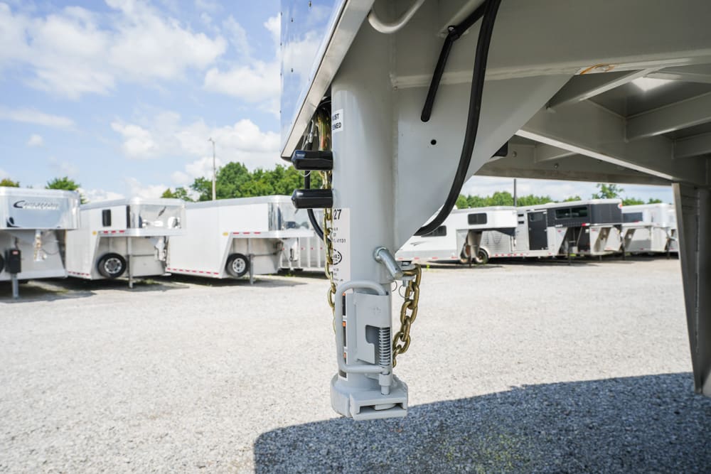 Close-up of a gray trailer hitch with safety chains and a locking mechanism, attached to a fifth-wheel trailer in a gravel lot with other trailers in the background under a blue sky.