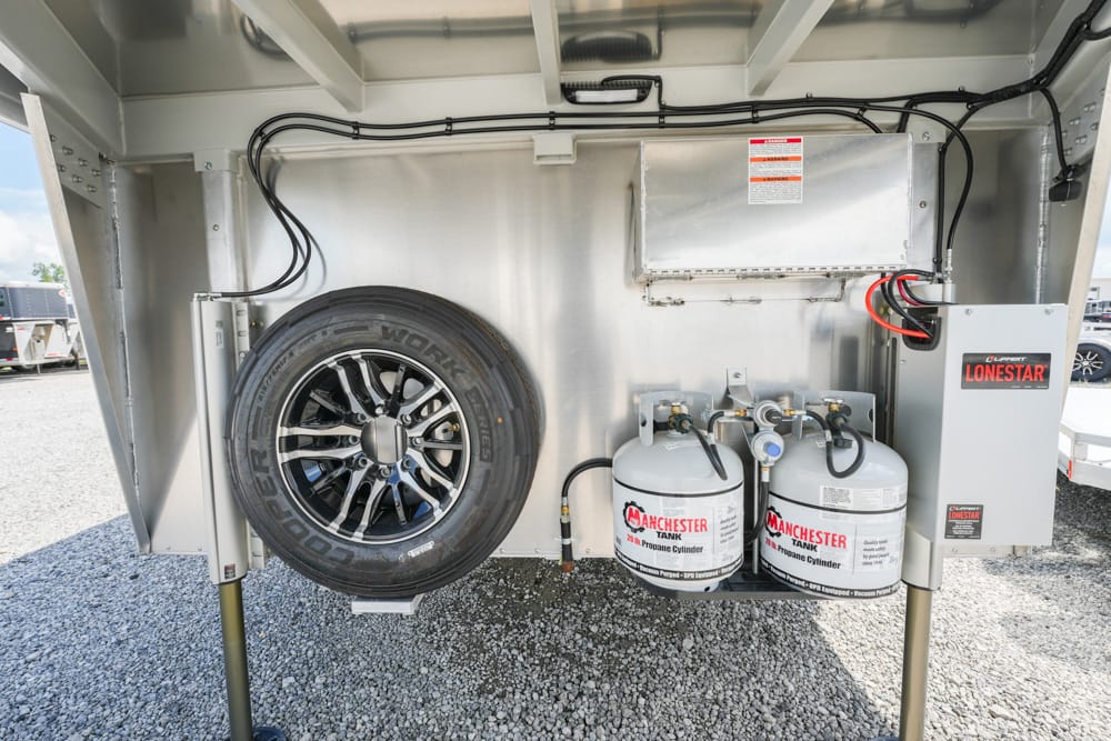 Side view of a utility trailer’s underside showing a mounted spare tire, two propane tanks, and an electrical control box/cabinet.