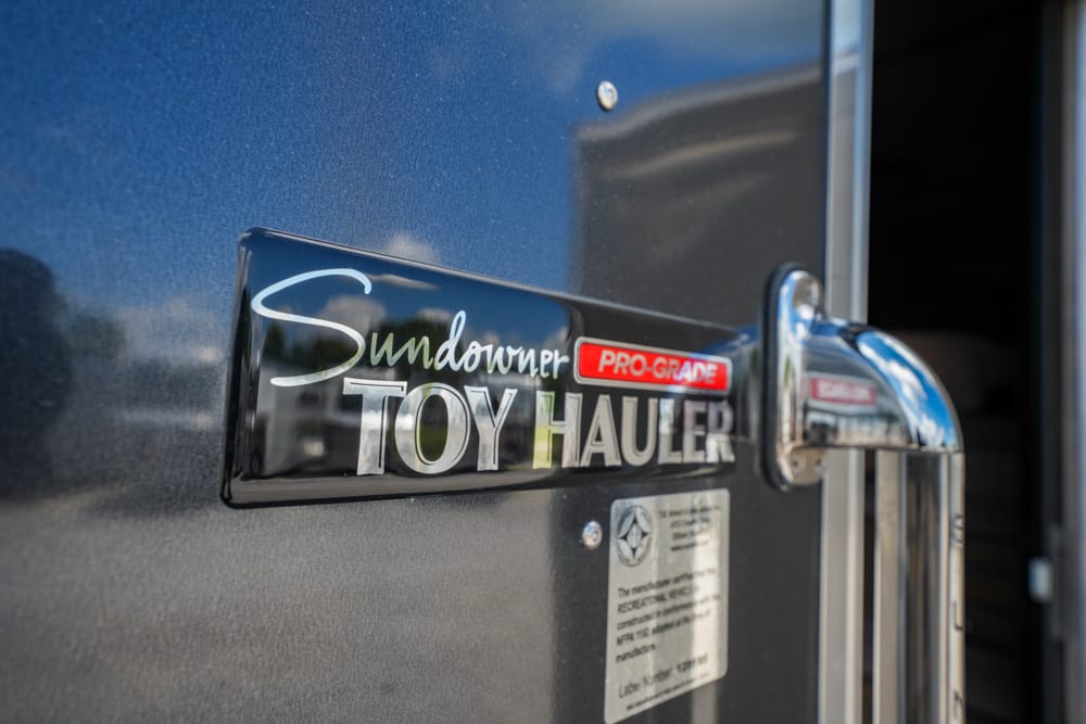 Badge on a dark Sundowner Toy Hauler trailer reading 'Sundowner PRO-GRADE TOY HAULER' with a chrome latch nearby