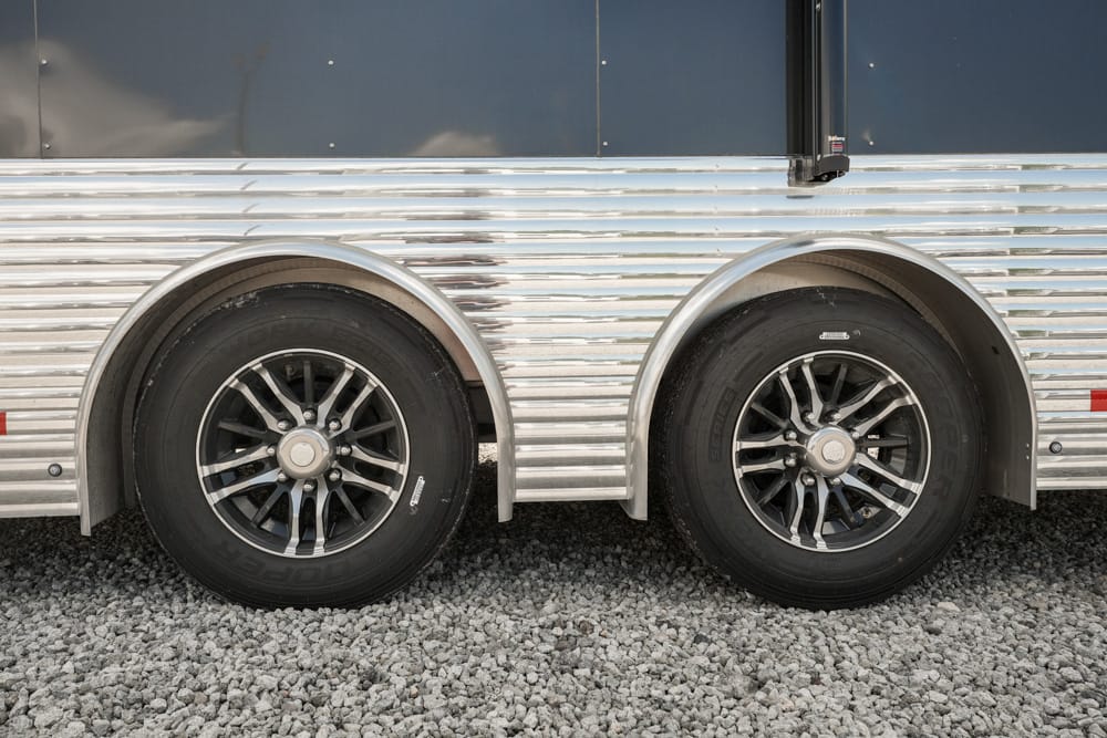 Two black tires with silver rims on a chrome trailer, parked on gravel ground.