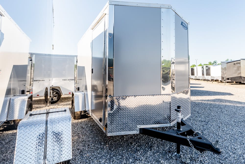 Silver enclosed cargo trailer with diamond-plate front near a hitch, parked on gravel among other trailers in a lot.