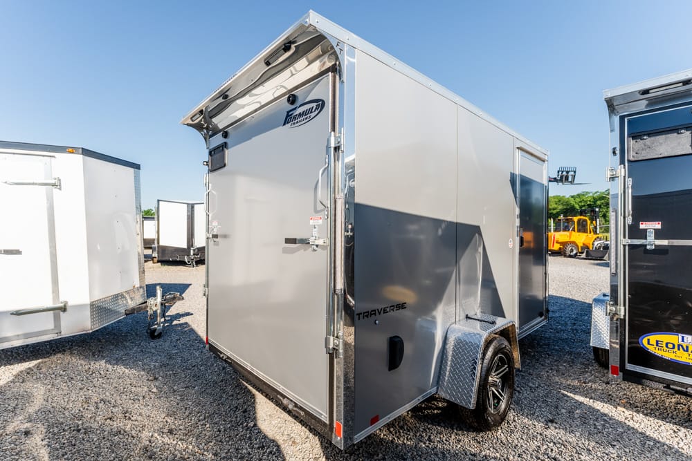Silver enclosed cargo trailer with black trim and diamond-plate fender, parked on gravel among other trailers under a clear blue sky.