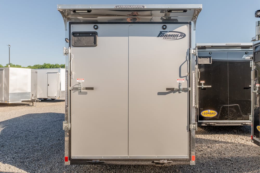 Rear view of a gray enclosed cargo trailer with double doors, latches, and a Formula Trailers logo on a gravel lot.