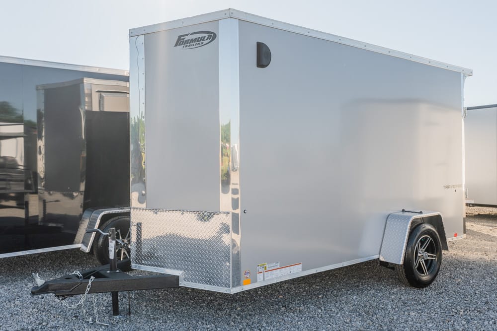 Silver enclosed cargo trailer with diamond-plate front, hitch and chains at the tongue, and a single black-wheeled axle on a gravel lot.