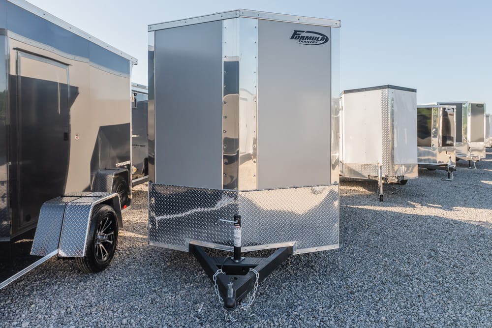 Row of enclosed cargo trailers on a gravel lot, with the closest gray trailer featuring a diamond-plate front and A-frame hitch.