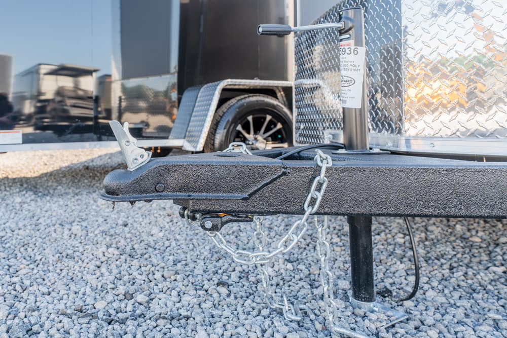Close-up of a trailer hitch with safety chains attached to a black frame on a gravel lot by a diamond-plate trailer side leg.