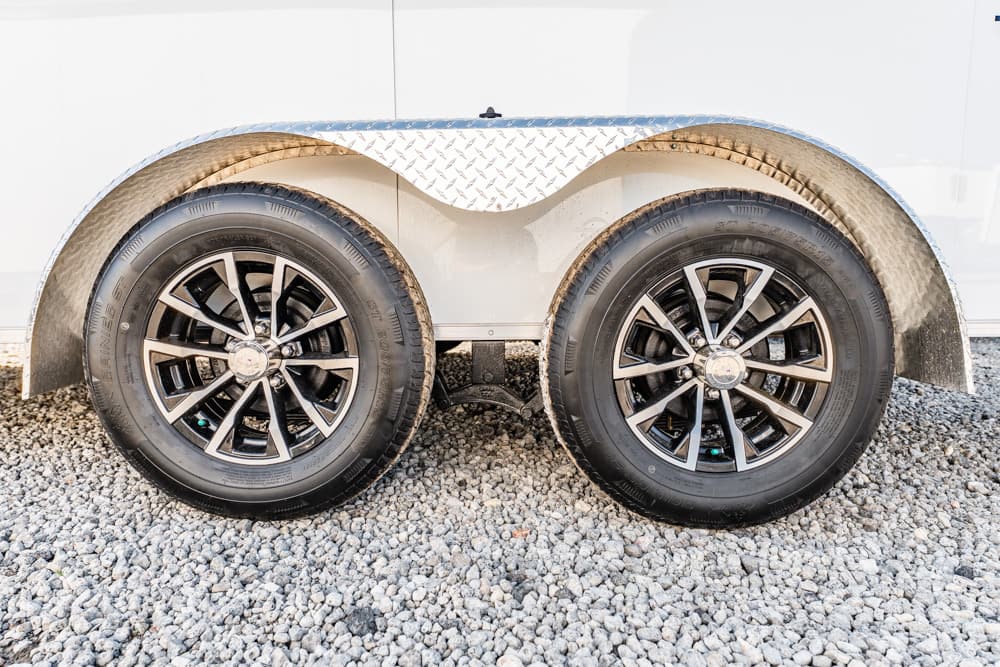 Dual trailer wheels with black and silver rims under a diamond-plate fender on a gravel surface