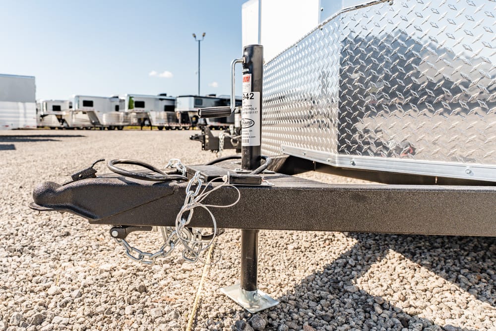 Close-up of a trailer hitch with safety chains, jack, and a diamond-plate trailer in a gravel RV lot.
