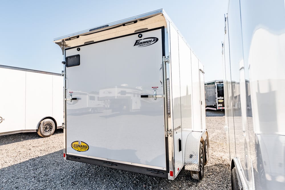 Rear of a white enclosed cargo trailer with a Formula trailer logo and a Leonard trailer sticker, parked in a gravel lot with other trailers nearby.