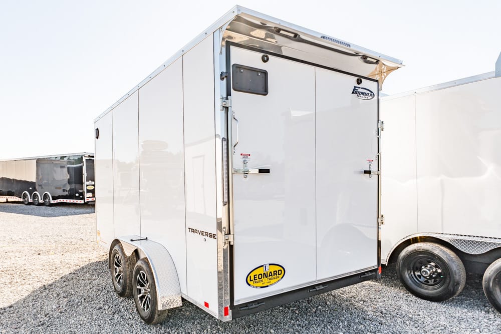 Rear view of a white enclosed cargo trailer with double back doors and a Leonard logo on the lower door panel, parked on gravel.