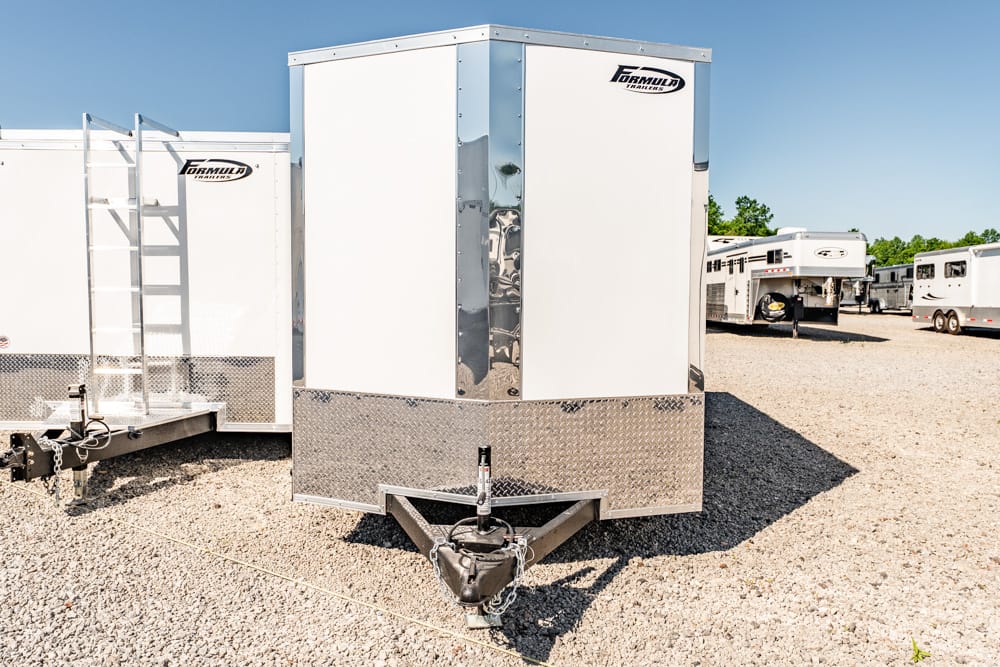 Front view of a white enclosed trailer with a diamond-plate rock guard and hitch, parked in a gravel lot among other trailers; clear blue sky overhead.