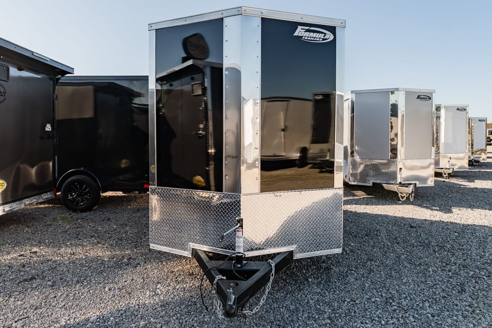 Front view of a silver enclosed cargo trailer with a diamond-plate front on a gravel lot, other trailers in the background.