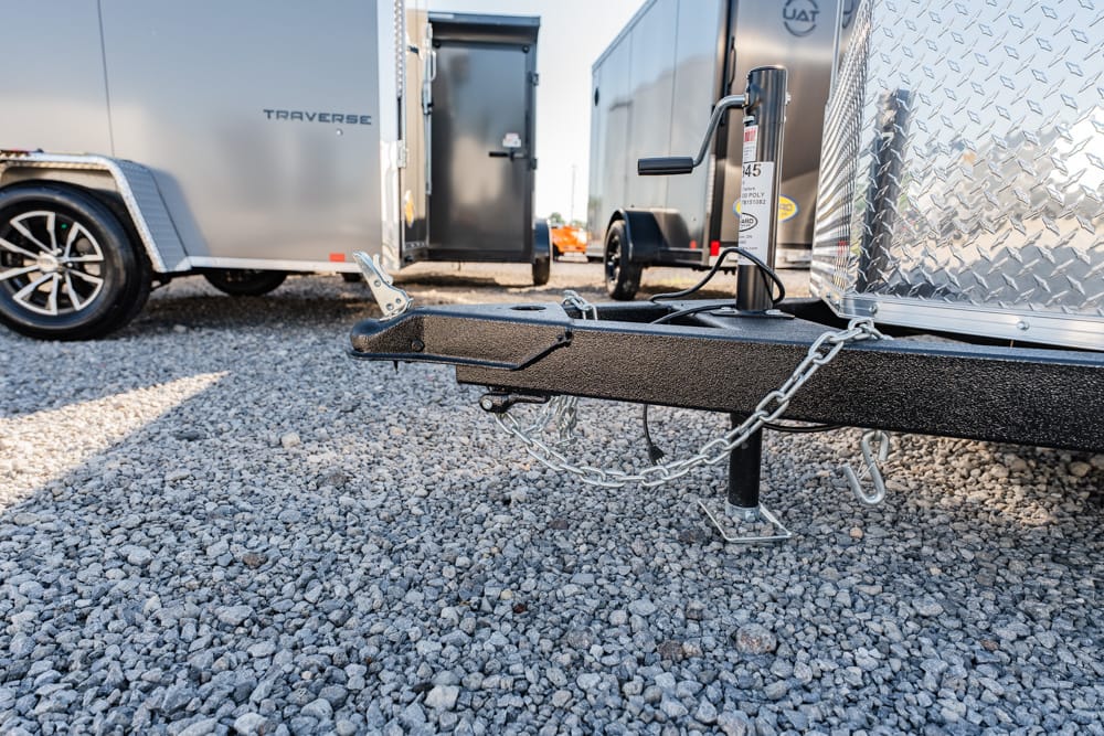 Close-up of a trailer hitch with safety chains and jack on a gravel lot, with other trailers in the background.