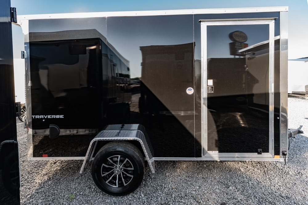 Black enclosed trailer with a steel diamond-plate fender and single wheel on gravel; door and windowless side visible, reflecting nearby objects.