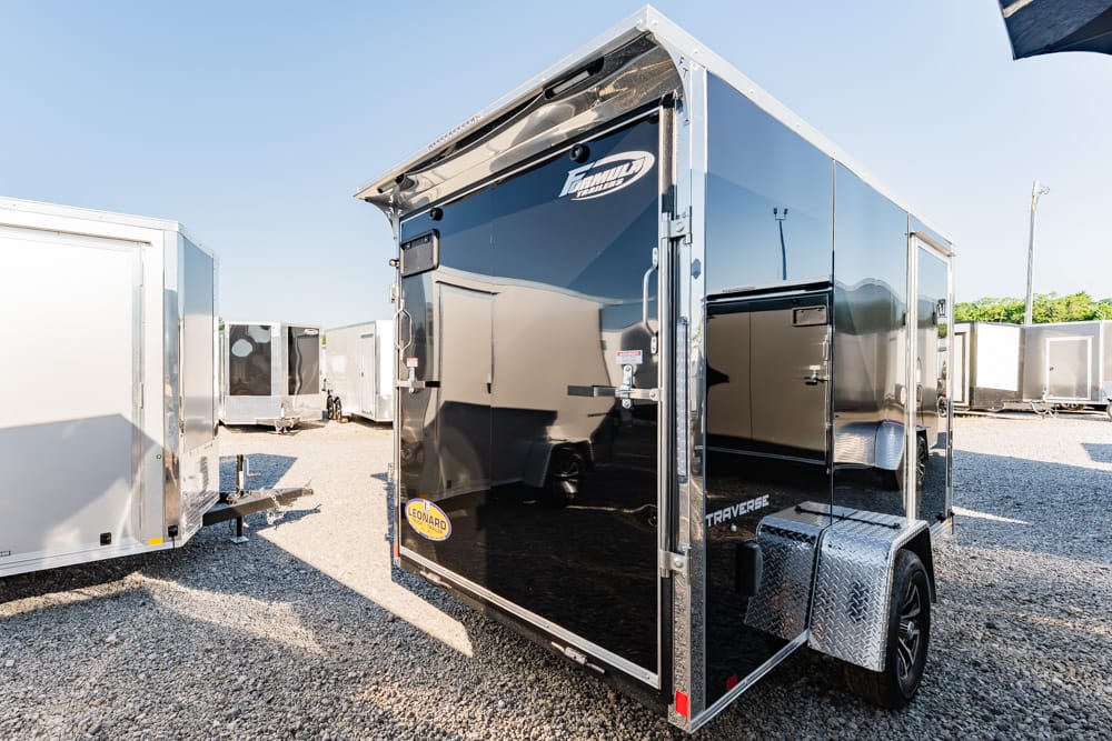 Black enclosed cargo trailer with a diamond-plate fender, parked on a gravel lot among other trailers; reflects surroundings in its glossy surface.
