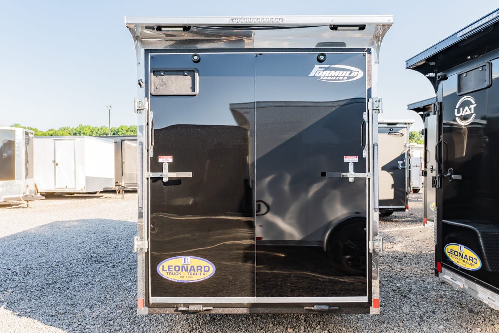 Rear view of a glossy black enclosed trailer with Leonard logo on the back doors, parked on a gravel lot with other trailers nearby.