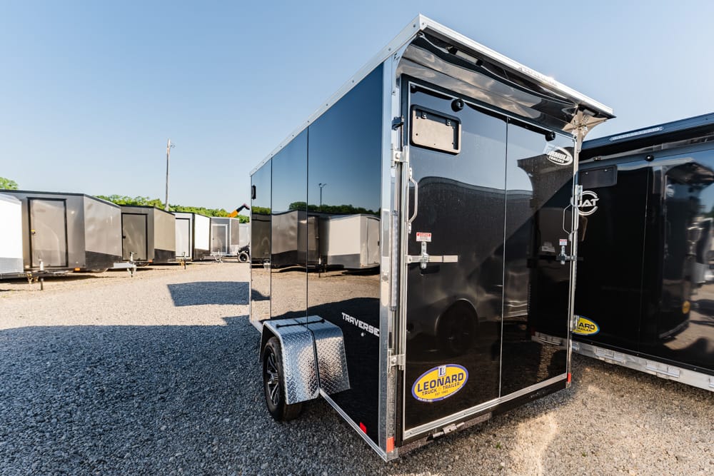 Black glossy enclosed cargo trailer with double rear doors and a diamond-plate fender, parked on gravel with other trailers in the background in a lot.