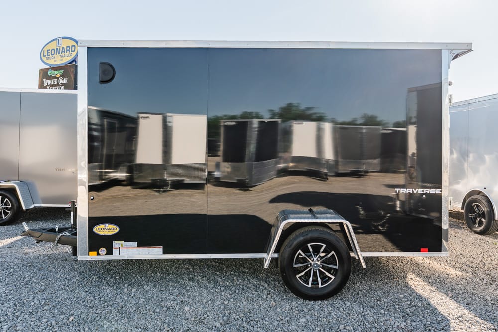 Black enclosed cargo trailer with a single axle on a gravel lot; reflects surrounding trailers in polished side panels, Traverse branding visible on rear panel, Leonard sign at front corner.