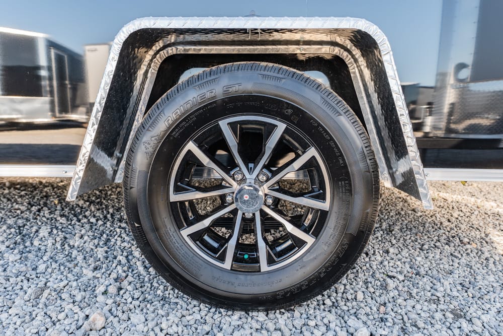 Close-up of a trailer wheel with a black alloy rim and a diamond-plate fender above a gravel surface.
