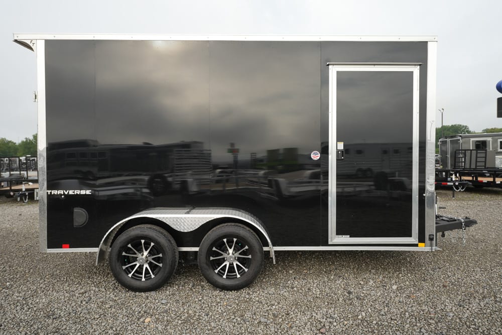 Black enclosed tandem-axle cargo trailer with a side entry door, parked on a gravel lot; reflections in the glossy surface show other trailers nearby.