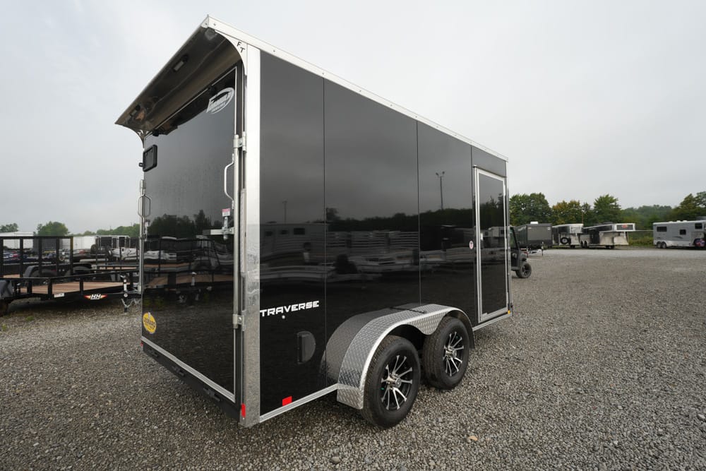 Black enclosed cargo trailer with dual axles and diamond-plate fenders in a gravel lot, with other trailers in the background.