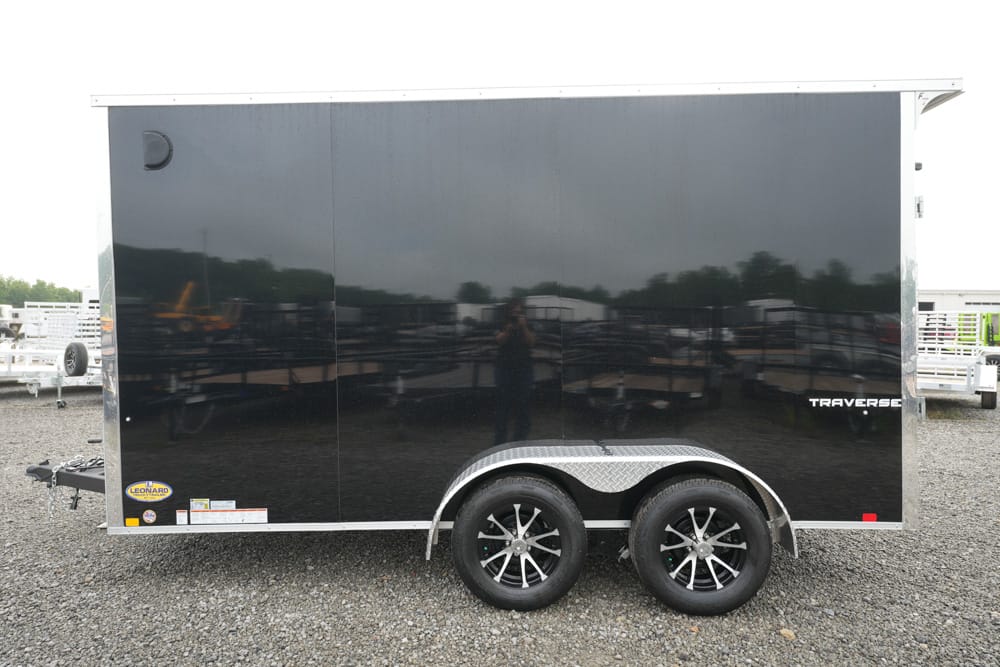 Black enclosed cargo trailer with two axles and diamond-plate fenders, parked on a gravel lot with other trailers in the background