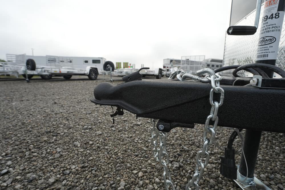 Close-up of a black trailer hitch with metal safety chains on a gravel lot, with trailers in the background.