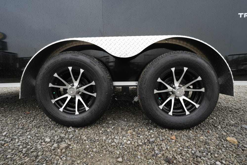 Dual trailer wheels with black tires and silver spokes under a diamond-plate fender on gravel ground