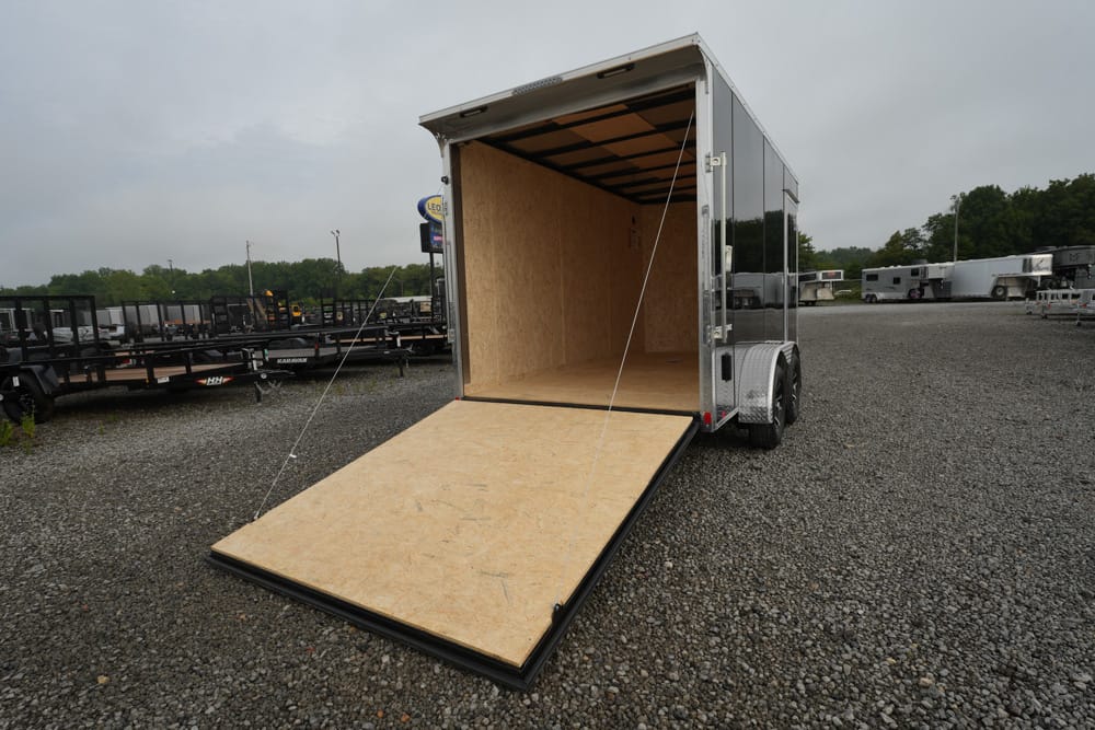Open cargo trailer with a plywood ramp down to a gravel lot, empty interior, trailer yard in the background