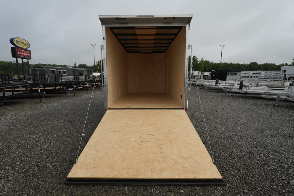 Open empty cargo trailer with ramp down, parked on gravel lot among other trailers and equipment at a sales lot.
