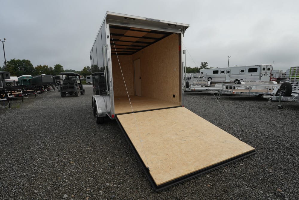 Open aluminum cargo trailer with its plywood interior and ramp deployed on a gravel lot, surrounded by other trailers and equipment.