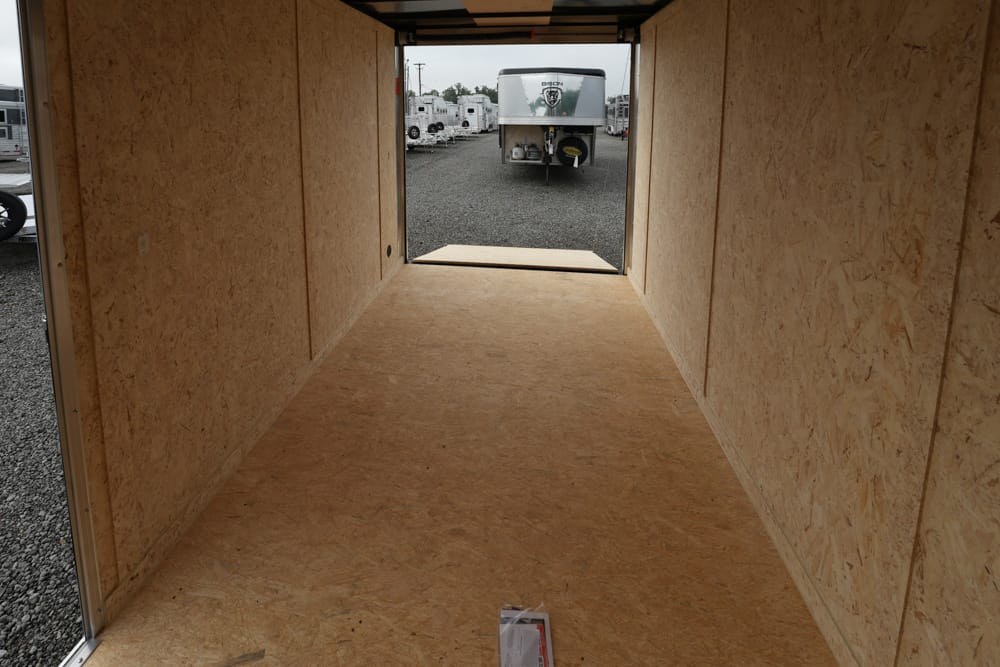 Empty plywood-walled trailer interior, looking toward the open rear with a gravel lot and parked trailers outside at the end of the aisle.