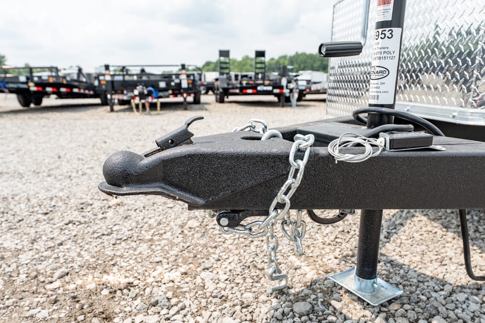 Close-up of a trailer tongue with a black coupler, safety chains, and a lock on a gravel lot, other trailers in the background