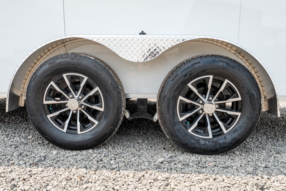 Dual-axle trailer wheels with black tires and silver spokes on a gravel surface under a white trailer, with a diamond-plate fender above.