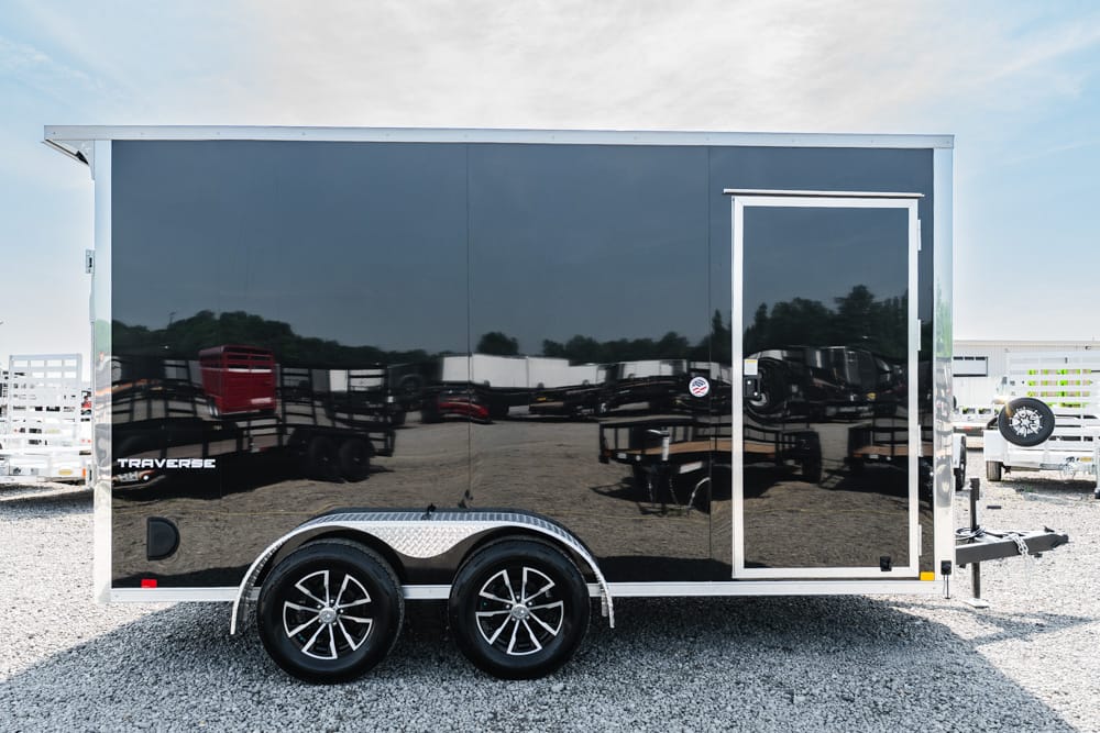 Black enclosed cargo trailer with dual axles, parked on gravel with a front hitch visible and Diamond Plate fenders on the wheel wells.
