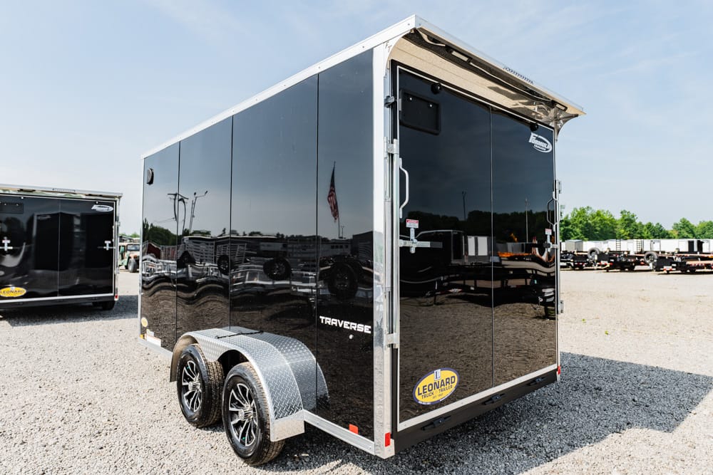 Black two-axle enclosed cargo trailer with diamond-plate fenders, parked on a gravel lot in bright sunlight.
