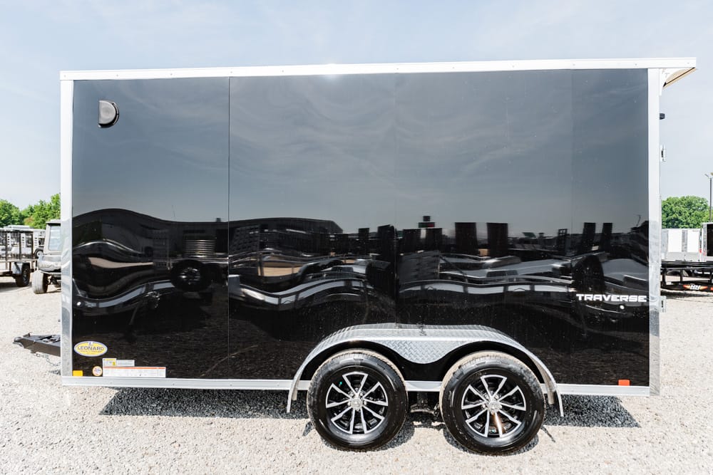 Glossy black enclosed two-axle cargo trailer with diamond-plate wheel wells on a gravel lot, reflections on its surface.