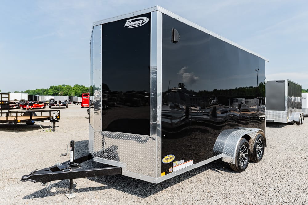 Black enclosed cargo trailer with diamond plate front, tandem aluminum wheels, and front hitch in a gravel lot.
