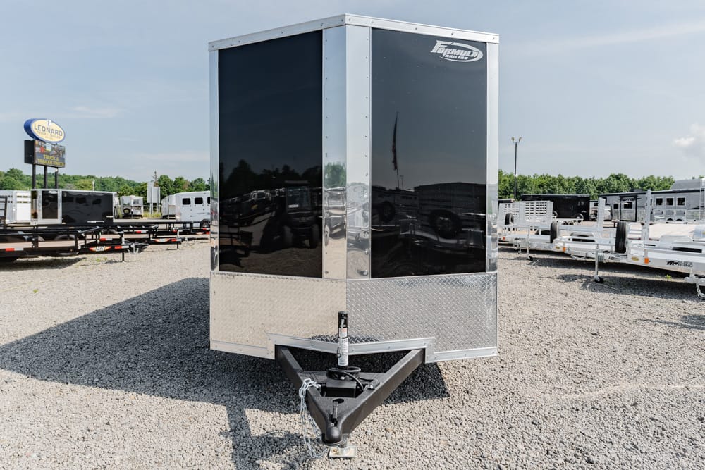 Front view of a black enclosed cargo trailer with a diamond-plate lower front, parked in a gravel trailer lot with other trailers in the background and a hitch at the front.