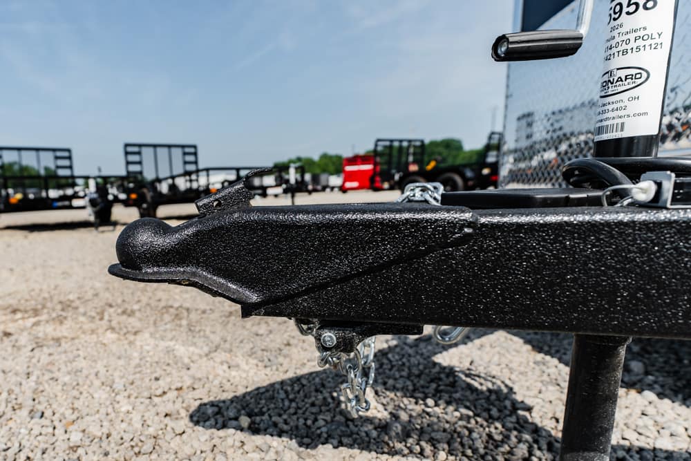 Close-up of a black trailer hitch on a gravel lot with other trailers in the background, silver chains visible nearby.