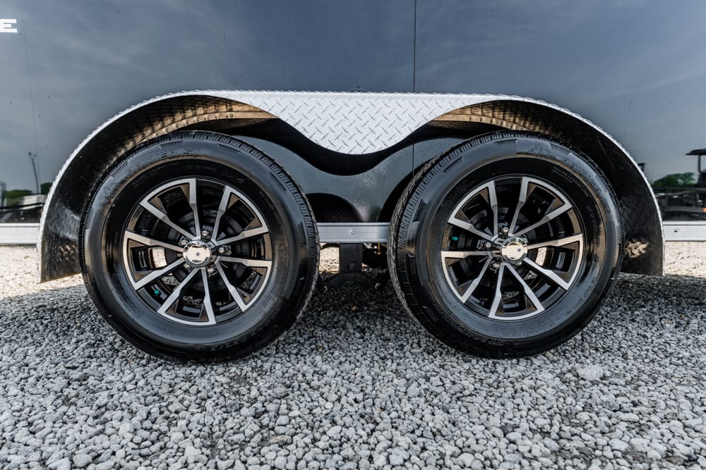 Dual-axle trailer with black alloy wheels and diamond-plate fender, parked on gravel.