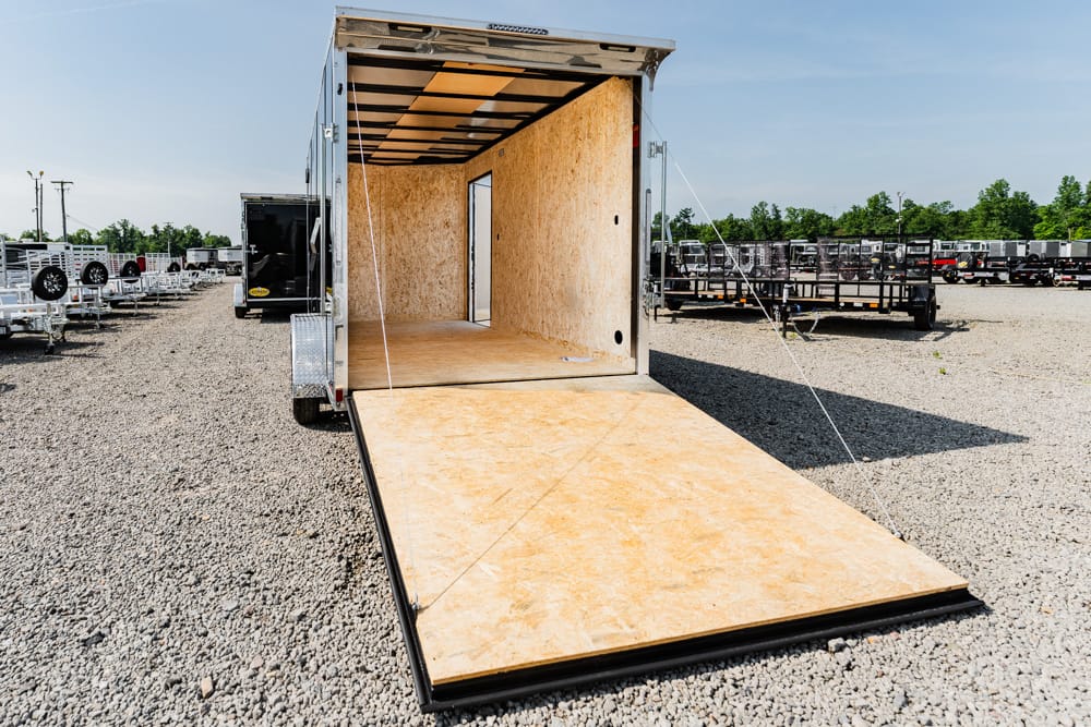 Open cargo trailer with ramp lowered, exposing plywood interior for loading in a gravel lot ready to move goods.