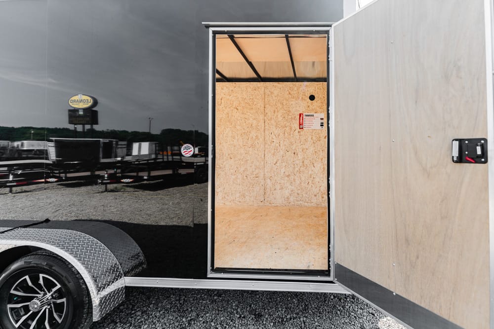 Open rear of a black enclosed trailer; unfinished plywood interior visible through the door, with a diamond‑plate wheel arch in the foreground.