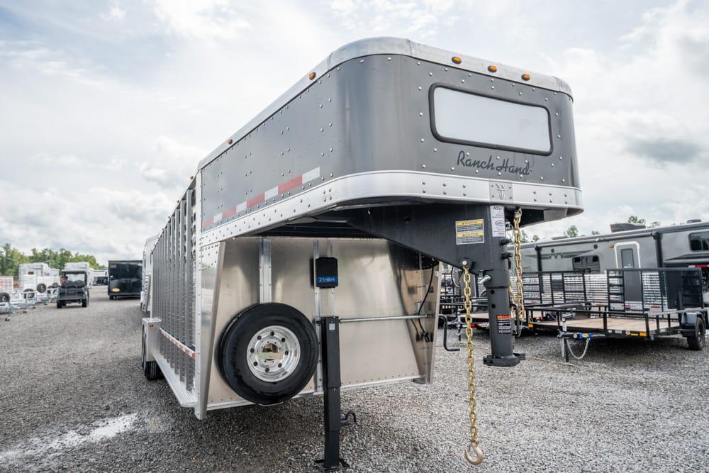 Silver aluminum gooseneck livestock trailer with spare tire mounted on side, parked on a gravel lot beside other trailers.