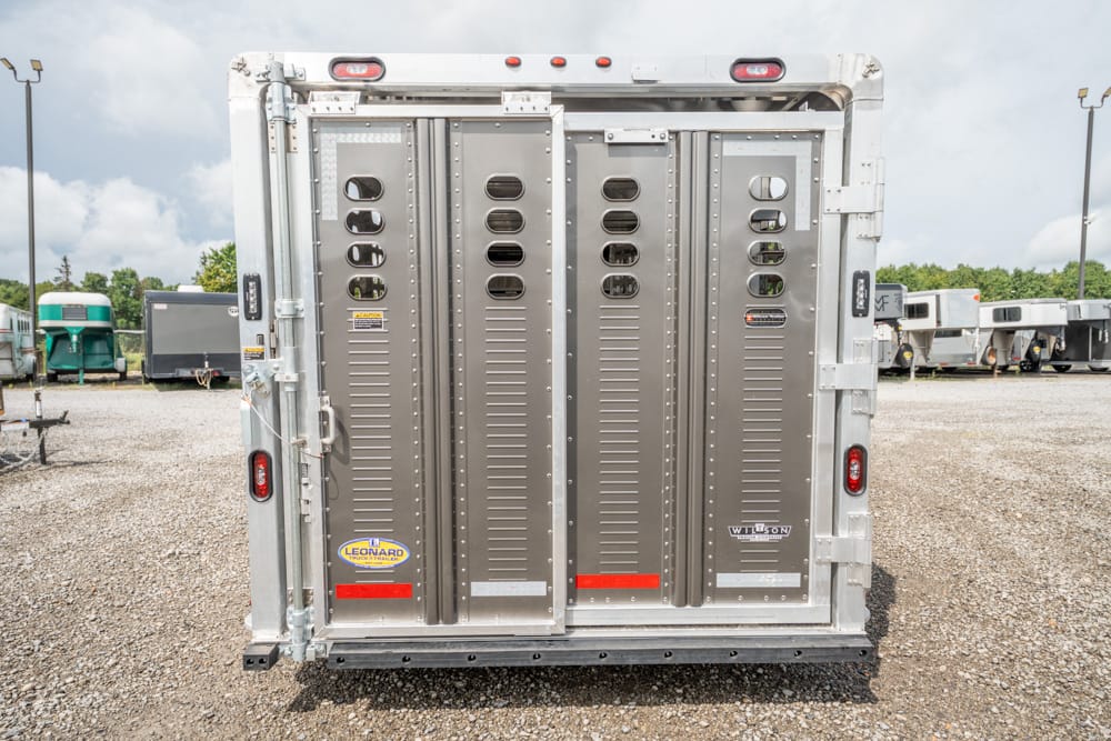 Rear view of a silver livestock trailer with two vertical doors, vents, and safety latches in a gravel lot. The trailer shows brand stickers and red reflectors.